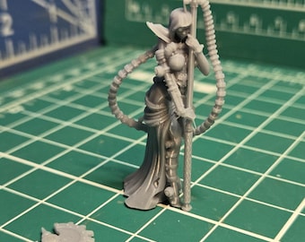 May include: A gray resin miniature figure of a hooded character standing and holding a tall staff with attached cables, accompanied by a separate small detached component piece, positioned on a green cutting mat with visible white grid lines.