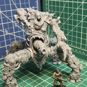 May include: A gray unpainted resin miniature of a multi-legged creature with a central maw and a cannon-like protrusion, standing next to a smaller painted soldier miniature on a green cutting mat with visible inch markings for scale.