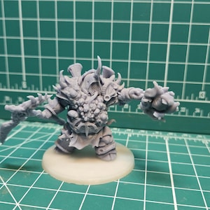 May include: A gray, unpainted miniature figure of a hulking, armored warrior with a spiked helmet, a cluster of organic growths on the chest, and a large, jagged weapon held in one hand, standing on a circular base.