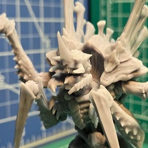 Puede incluir: Una figura en miniatura gris claro de una criatura con múltiples extremidades y una cabeza puntiaguda. La figura está posada sobre una superficie de cuadrícula verde y azul, lo que sugiere un juego de mesa o un artículo de hobby.