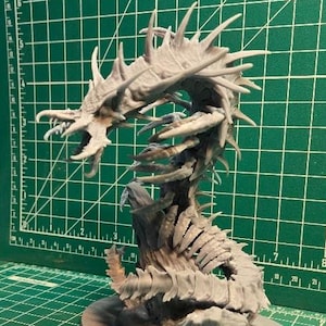 May include: A grey, unpainted resin miniature of a serpentine creature with multiple spikes and a skeletal body structure, positioned next to a smaller, painted gold and green armoured soldier miniature on a green cutting mat with grid lines.