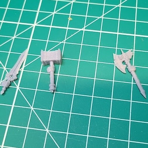 May include: Four miniature plastic weapons, including a sword, a hammer, an axe, and a dagger, on a green grid surface.