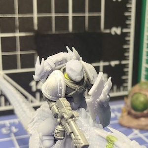 May include: A light gray unpainted miniature figure of a warrior riding a reptilian creature. The warrior wears armor and holds a rifle. A small brown and green spherical object and a gray wall segment are nearby on a blue cutting mat.