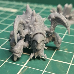May include: A set of three unpainted grey resin miniature figures featuring a central creature with sharp claws and spikes, a smaller rounded creature to the left, and a crystalline cluster to the right, all displayed on a green cutting mat.