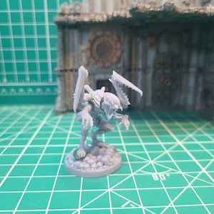 May include: A light gray unpainted miniature figure of a creature with sharp, blade-like appendages on its back and arms. The figure is in a crouching position on a circular base textured with small skulls, featuring a small green ring on its shoulder.