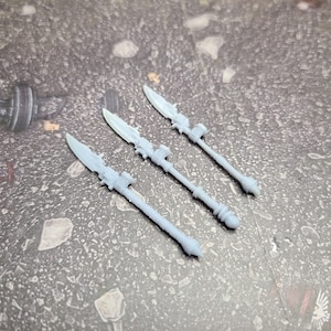 May include: Four white plastic miniature spears with a pointed tip and a decorative handle. The spears are arranged in a row on a gray textured surface.