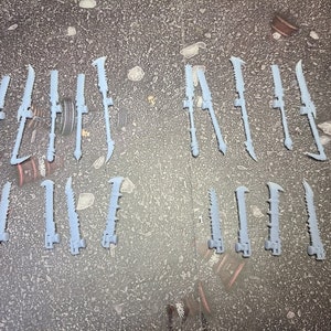 Puede incluir: Una colección de dieciséis armas de plástico en miniatura de color gris claro, cada una con una hoja larga y dentada unida a un mango con una base circular. Estos pequeños componentes detallados están dispuestos en dos filas, mostrando un diseño uniforme.