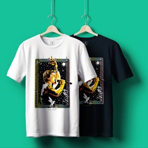 May include: Two t-shirts with a colorful graphic print of a person singing into a microphone. The graphic is surrounded by a colorful frame with stars and a sun. The text "SHOUT LOUDER" is at the bottom of the graphic. One t-shirt is white and the other is black.