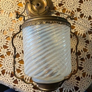 May include: Antique brass-toned wall sconce with a cylindrical, swirled glass shade. The shade is white and translucent, with a spiral pattern. The sconce has decorative metalwork and a mounting plate.