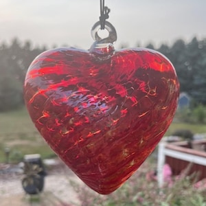 May include: A hanging, heart-shaped ornament made of red glass. The glass has a textured appearance with streaks of lighter red and gold. The ornament is suspended by a black string and a clear glass loop.
