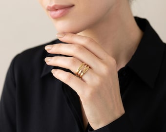 May include: A set of three stacked gold-toned rings worn on a finger. Each ring features a smooth, polished finish and is engraved with a distinct name in a clear, sans-serif font. The rings are thin, circular bands that sit flush against each other.