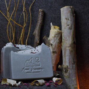 May include: A gray bar of soap with a mountain and moon design and the text "Anya Handmade" on a gray surface with birch bark and twigs.