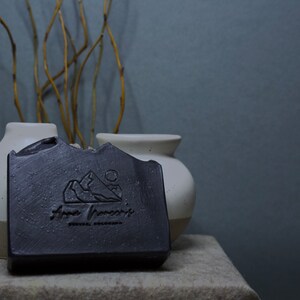 May include: A dark gray bar of soap with a mountain and sun logo, and the text "Aqua Francesca, Denver, Colorado". The soap is placed in front of two white vases and some decorative branches.