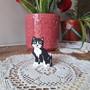 May include: A black and white cat figurine with green eyes sits on a white lace doily. The cat is painted with a tuxedo pattern. A pink flower pot with red roses and a green plant are in the background.