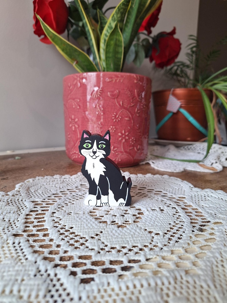 May include: A black and white cat figurine with green eyes sits on a white lace doily. The cat is painted with a tuxedo pattern. A pink flower pot with red roses and a green plant are in the background.