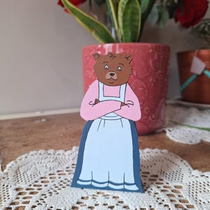 May include: A wooden bear figurine wearing a pink top and a light blue skirt with a dark blue trim. The bear has brown fur and is standing on a white lace doily. A red flower pot is in the background.