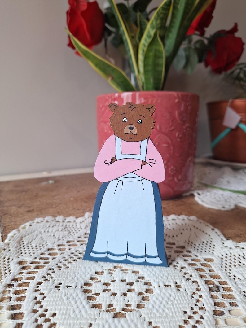 May include: A wooden bear figurine wearing a pink top and a light blue skirt with a dark blue trim. The bear has brown fur and is standing on a white lace doily. A red flower pot is in the background.
