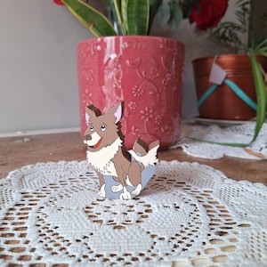May include: A wooden dog figurine with brown and white fur, a red tongue, and a blue base. The dog is standing on a white lace doily, with a pink flower pot and plants in the background.