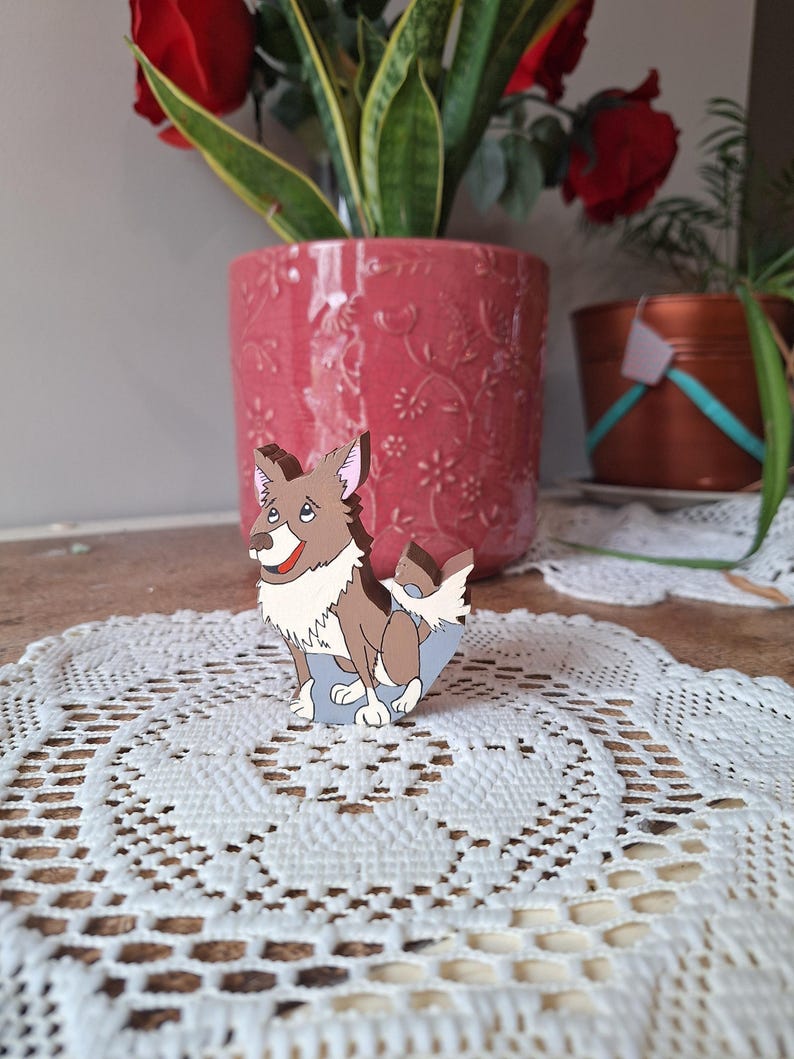 May include: A wooden dog figurine with brown and white fur, a red tongue, and a blue base. The dog is standing on a white lace doily, with a pink flower pot and plants in the background.