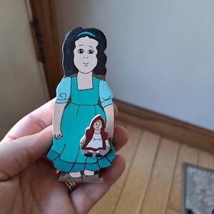 May include: A hand-painted wooden figure of a woman in a teal dress holding a smaller figure of a girl in a red hooded cape. The larger figure has black hair and a teal headband. The figures are on a wooden base.