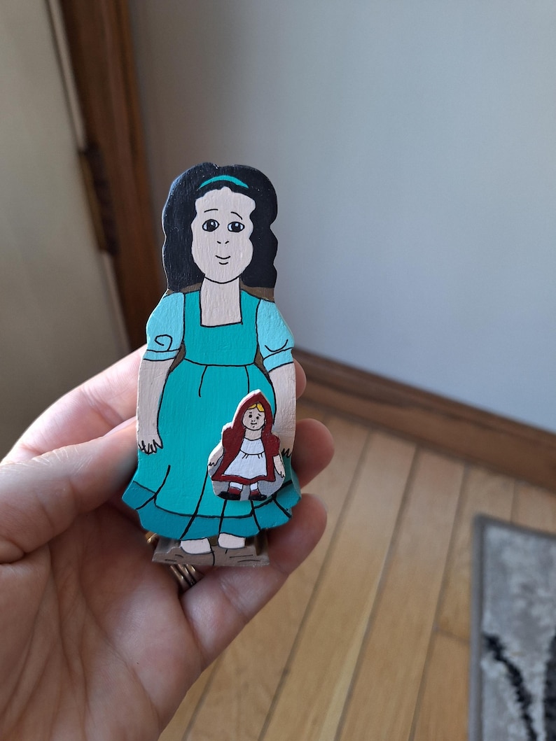 May include: A hand-painted wooden figure of a woman in a teal dress holding a smaller figure of a girl in a red hooded cape. The larger figure has black hair and a teal headband. The figures are on a wooden base.