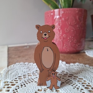May include: Two wooden bear figurines. The larger bear is brown with a lighter brown belly and a smiling face. A smaller bear figurine is in front of the larger bear. The background includes a pink flower pot and a white lace doily.