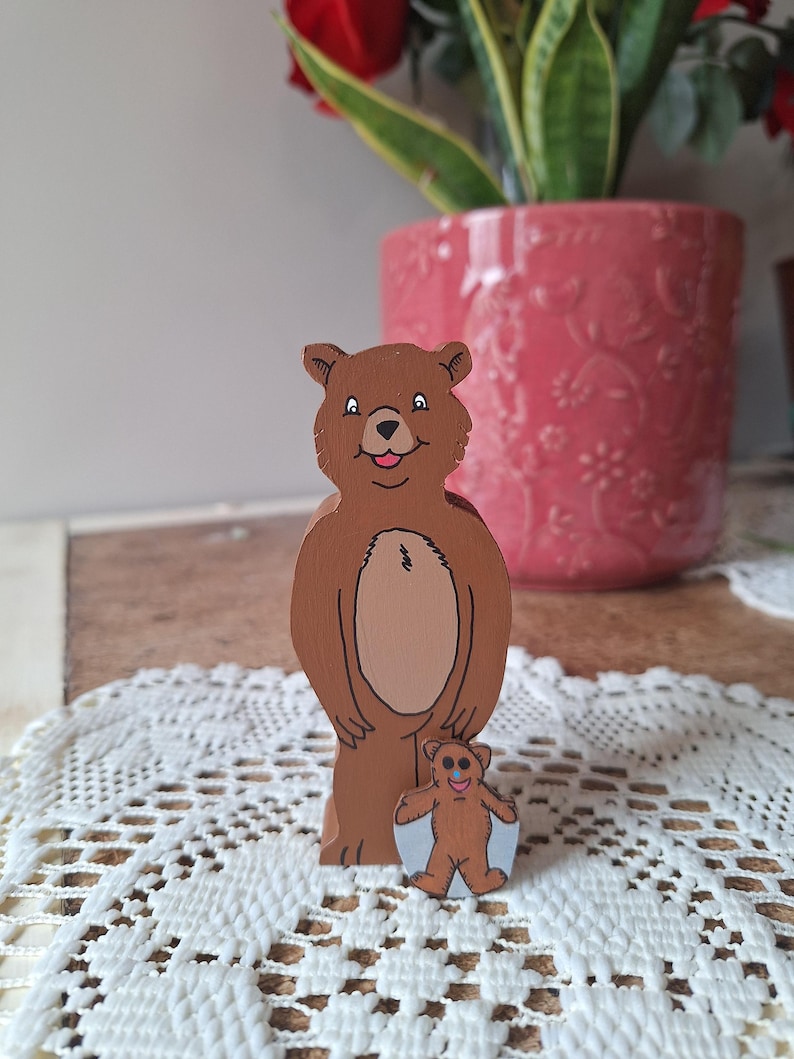 May include: Two wooden bear figurines. The larger bear is brown with a lighter brown belly and a smiling face. A smaller bear figurine is in front of the larger bear. The background includes a pink flower pot and a white lace doily.