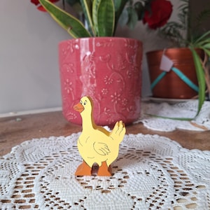 May include: A yellow wooden goose figurine with orange feet and beak. The goose is standing on a white lace doily. A pink flower pot with a floral design and a plant are in the background.
