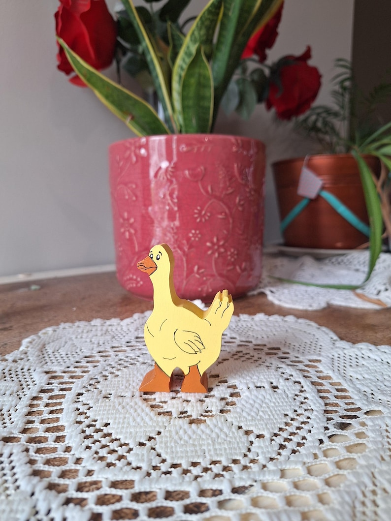 May include: A yellow wooden goose figurine with orange feet and beak. The goose is standing on a white lace doily. A pink flower pot with a floral design and a plant are in the background.