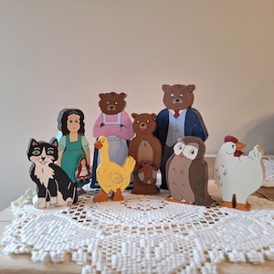 May include: A collection of wooden storybook characters, including a black and white cat, a girl in a green dress, a yellow duck, several bears, an owl, and a white chicken. The figures are painted with bright colors and stand on a white lace doily.