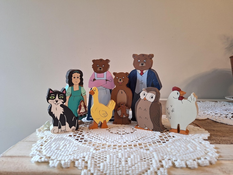 May include: A collection of wooden storybook characters, including a black and white cat, a girl in a green dress, a yellow duck, several bears, an owl, and a white chicken. The figures are painted with bright colors and stand on a white lace doily.