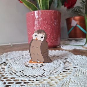 May include: A wooden owl figurine with a brown body, tan chest, and orange feet. The owl has large, round eyes and is perched on a white lace doily. A pink flower pot with red roses and a copper pot are in the background.