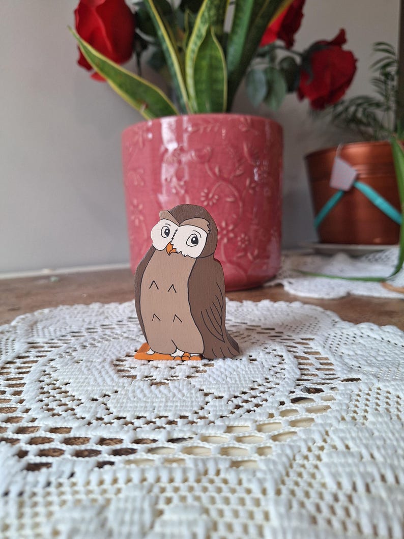 May include: A wooden owl figurine with a brown body, tan chest, and orange feet. The owl has large, round eyes and is perched on a white lace doily. A pink flower pot with red roses and a copper pot are in the background.