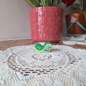 May include: A green and yellow snake-shaped pin rests on a white lace doily. The pin has a gray stripe and a smiling face. A pink floral pot and a copper pot are in the background.