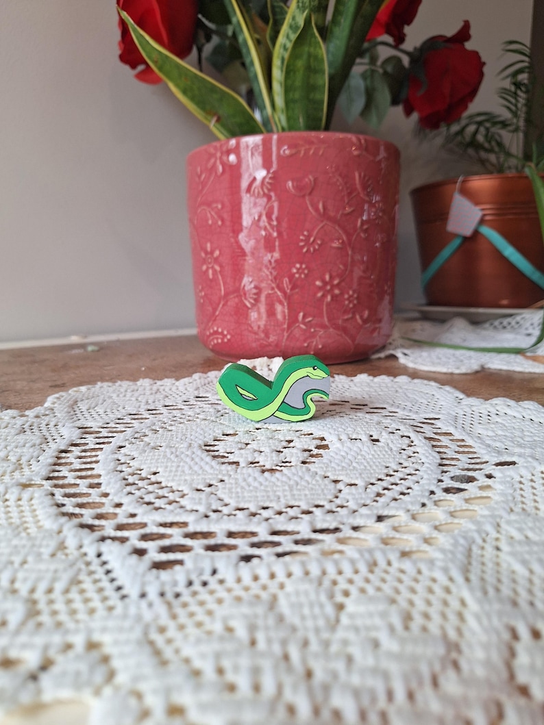 May include: A green and yellow snake-shaped pin rests on a white lace doily. The pin has a gray stripe and a smiling face. A pink floral pot and a copper pot are in the background.