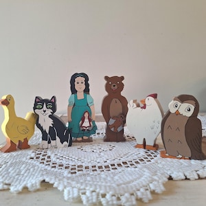 May include: A collection of colorful wooden storybook characters, including a yellow duck, a black and white cat, a girl in a teal dress, a brown bear, a white chicken, and a brown owl. The figures are standing on a white lace doily.