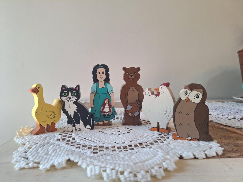 May include: A collection of colorful wooden storybook characters, including a yellow duck, a black and white cat, a girl in a teal dress, a brown bear, a white chicken, and a brown owl. The figures are standing on a white lace doily.
