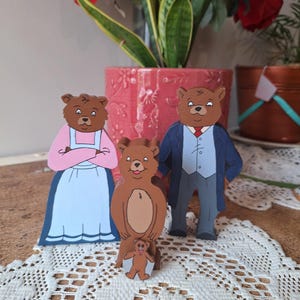 May include: Wooden bear family figurines. The figures include a mother bear in a pink dress and blue skirt, a father bear in a blue suit, and two smaller bear figures. The bears are brown with black details and are standing on a white lace tablecloth.