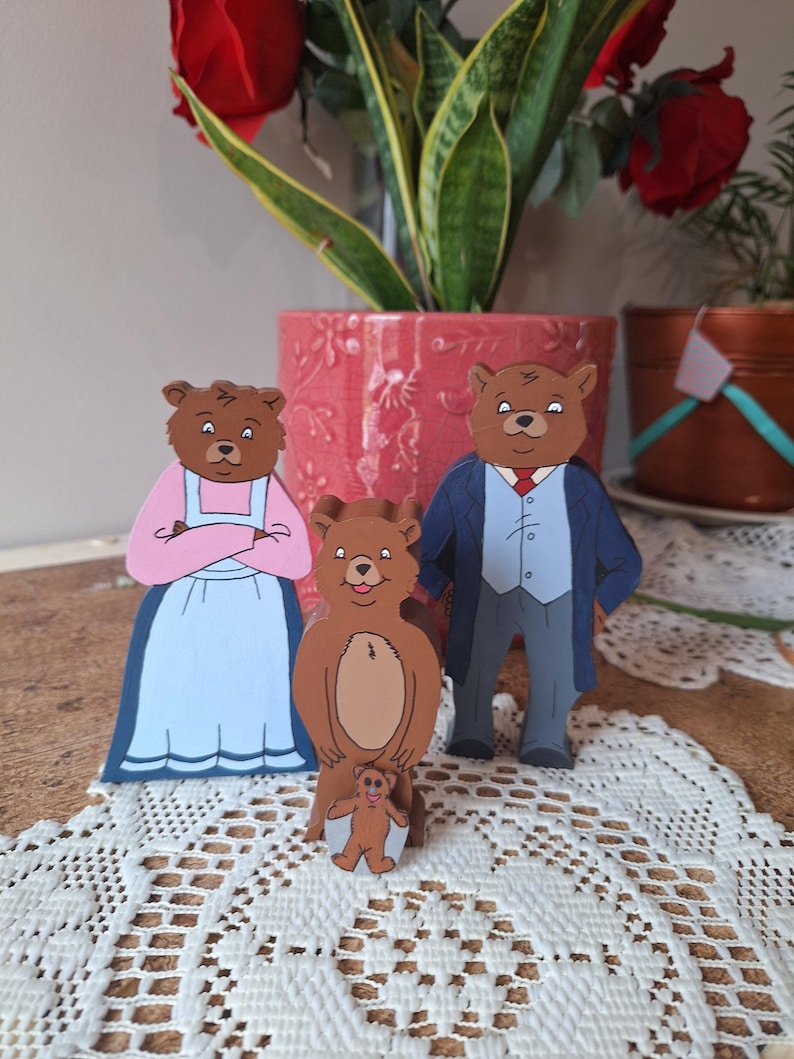May include: Wooden bear family figurines. The figures include a mother bear in a pink dress and blue skirt, a father bear in a blue suit, and two smaller bear figures. The bears are brown with black details and are standing on a white lace tablecloth.