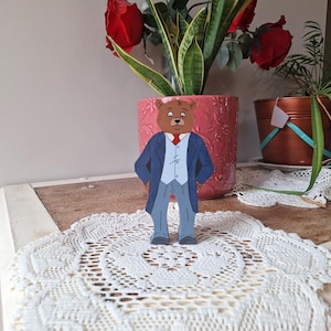 May include: A wooden bear figurine wearing a blue suit, gray pants, and a red tie stands on a white lace doily. The bear is in front of a pink flower pot with red roses and green plants.
