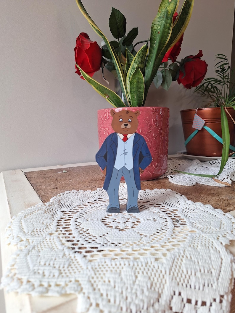 May include: A wooden bear figurine wearing a blue suit, gray pants, and a red tie stands on a white lace doily. The bear is in front of a pink flower pot with red roses and green plants.