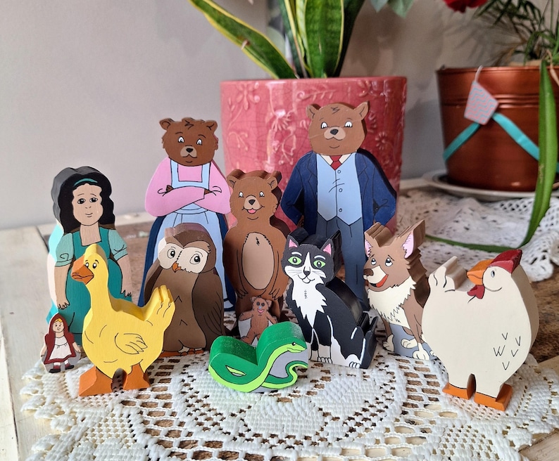 May include: A collection of wooden storybook characters, including a duck, owl, cat, bear, and a girl in a red cloak. The figures are brightly colored and stand on a white lace surface. The characters are from a children's story.
