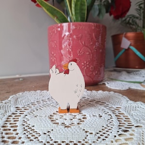 May include: A wooden chicken figurine with a white body, red comb, and orange feet stands on a white lace doily. The chicken has a black beak and black markings. A pink flower pot with red roses and green plants is in the background.