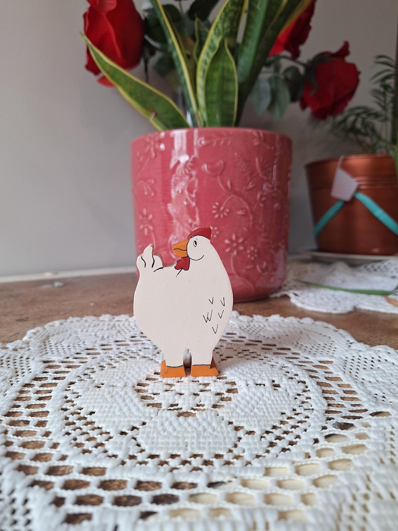 May include: A wooden chicken figurine with a white body, red comb, and orange feet stands on a white lace doily. The chicken has a black beak and black markings. A pink flower pot with red roses and green plants is in the background.