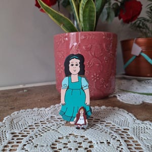 May include: Two wooden figurines depicting characters from a fairy tale. The larger figure wears a teal dress and has black hair. The smaller figure is dressed in a red hooded cloak. The figurines are placed on a white lace doily.