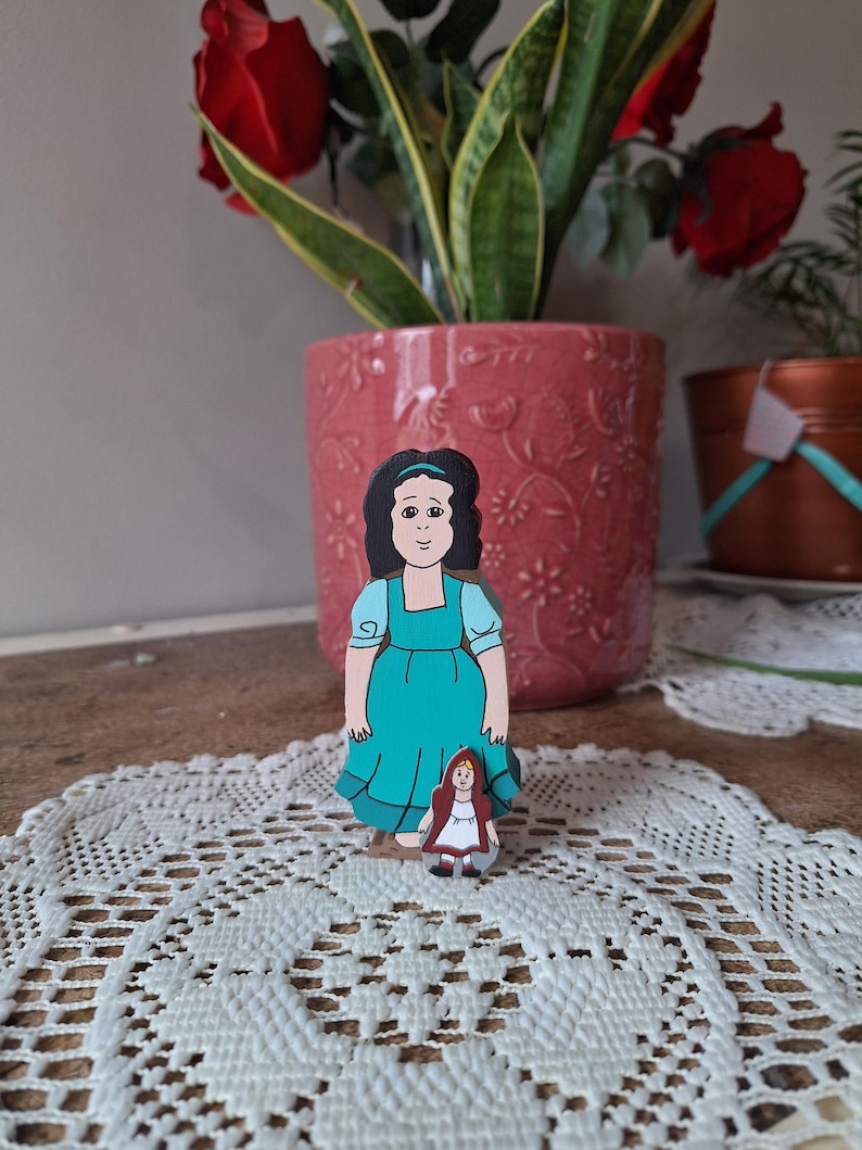 May include: Two wooden figurines depicting characters from a fairy tale. The larger figure wears a teal dress and has black hair. The smaller figure is dressed in a red hooded cloak. The figurines are placed on a white lace doily.