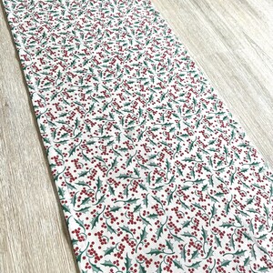 May include: White table runner with a repeating pattern of green holly leaves and red berries.