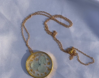 May include: A gold chain necklace with a round pendant featuring a pressed white flower with gold flecks inside.
