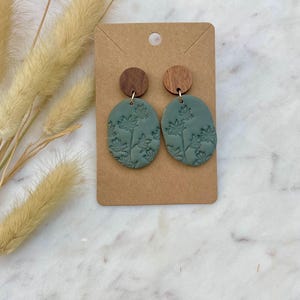Pode incluir: Um par de brincos de argila verde com um design floral e detalhes em madeira. Os brincos são de forma oval e têm uma superfície texturizada.