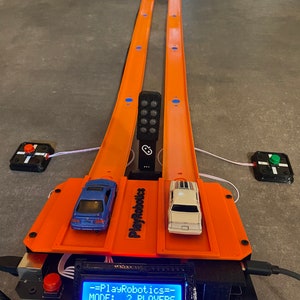 Könnte beinhalten: Eine zweispurige orangefarbene Rennstrecke mit zwei Spielzeugautos an der Startlinie. Die Strecke ist mit einem schwarzen Bedienfeld mit einem blauen LCD-Bildschirm verbunden, auf dem "PlayRobotics MODE: 2 PLAYERS" steht.